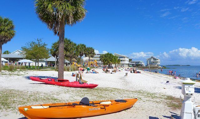 People relax on a sandy beach with palm trees, colorful kayaks, and nearby buildings. Some enjoy the sun while others play near the water under a bright blue sky with scattered clouds.