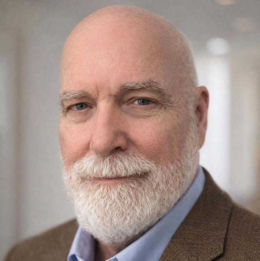 A bearded older man with a bald head, wearing a brown blazer and light blue shirt, smiles gently while looking at the camera against a softly blurred indoor background.