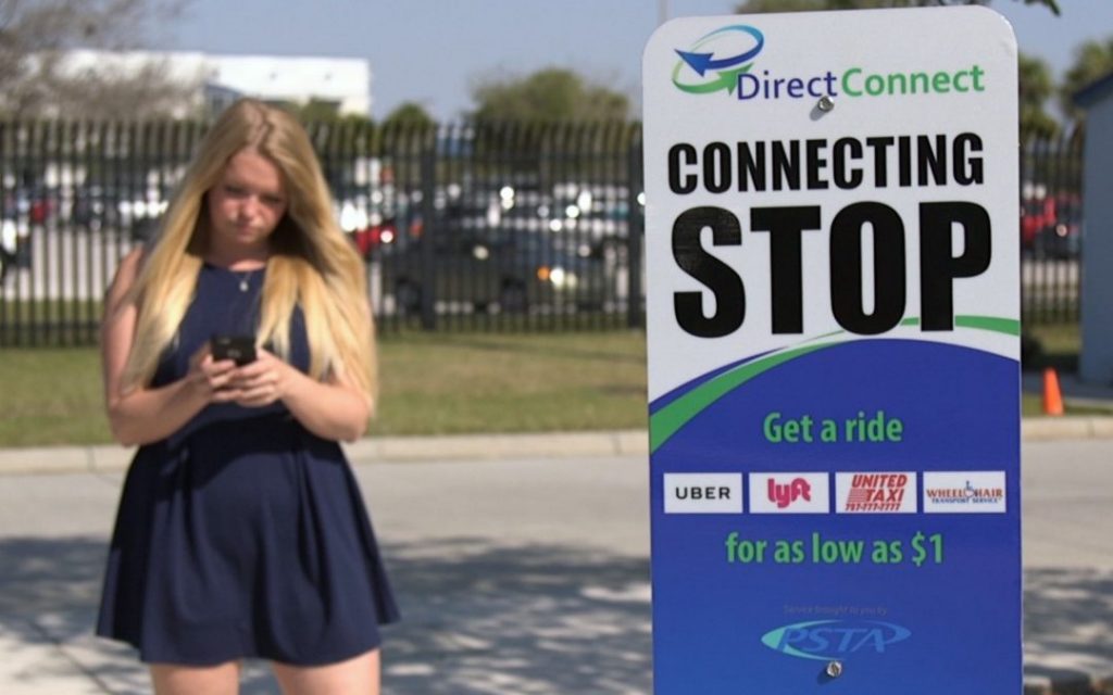A woman in a blue dress stands outdoors, looking at her phone next to a Direct Connect sign for a rideshare connecting stop, showing logos for Uber, Lyft, and others. Cars and a fence are visible in the background.
