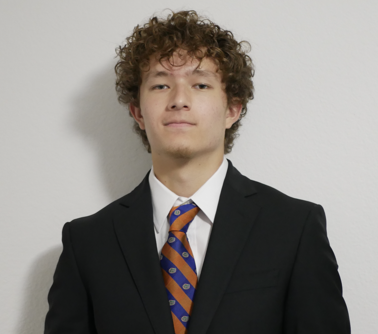 A young man with curly brown hair is wearing a black suit, white shirt, and a blue and orange patterned tie, standing against a plain white wall.