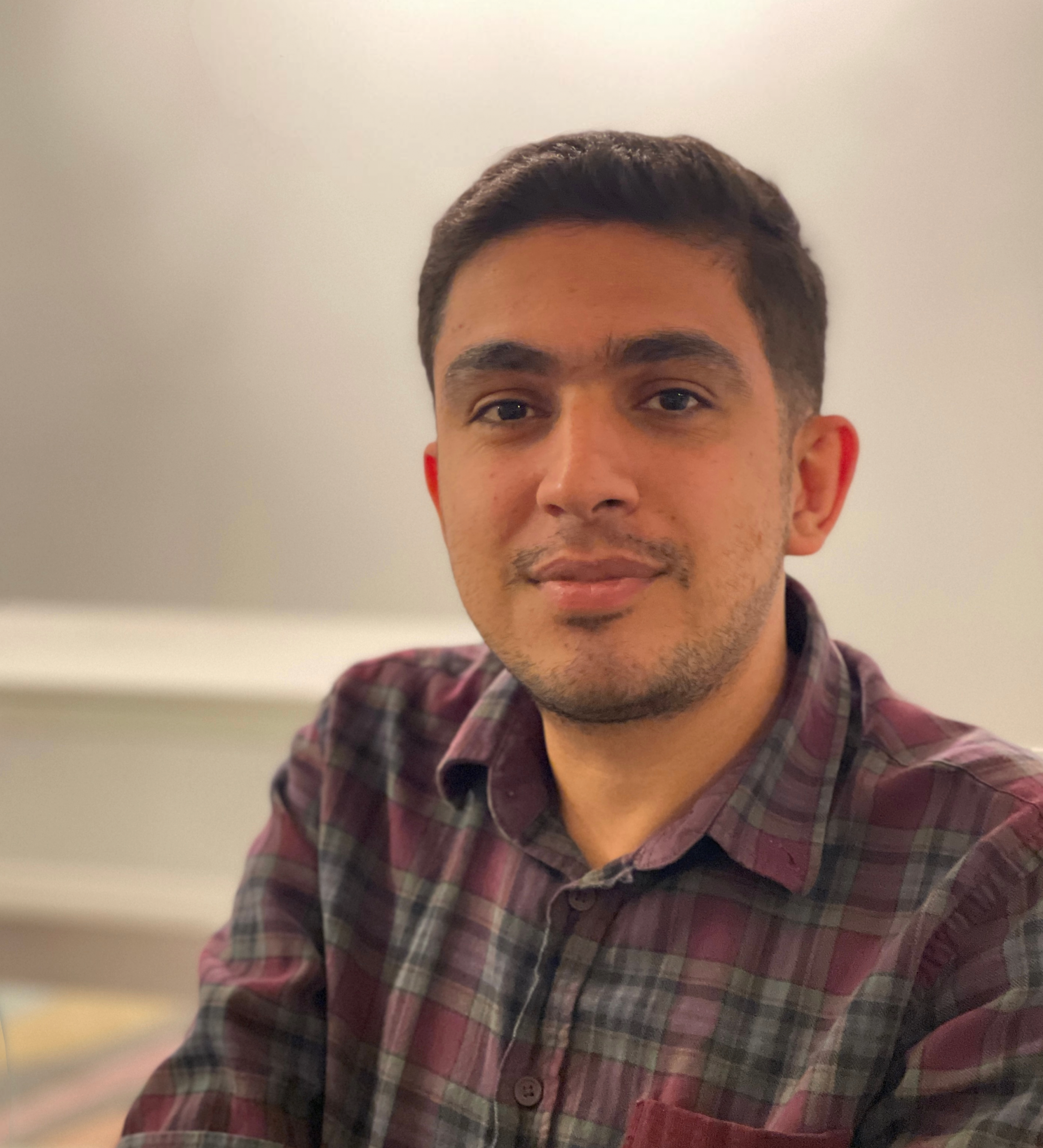 A man with short dark hair and a trimmed beard is wearing a maroon and gray plaid shirt, sitting indoors with a neutral background and looking at the camera with a slight smile.
