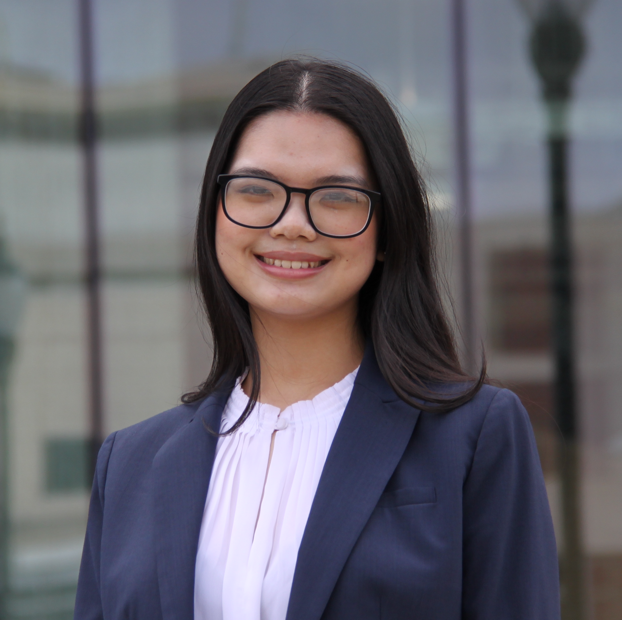 A young woman with long dark hair and glasses is smiling, wearing a navy blazer over a white blouse. She is standing in front of a glass building.