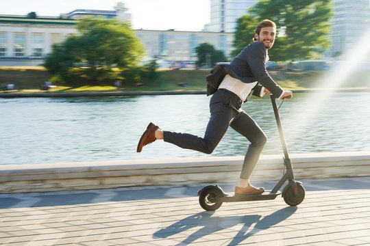 A man in a suit happily rides an electric scooter along a riverside path on a sunny day, with one foot pushing off the ground and buildings and trees in the background.