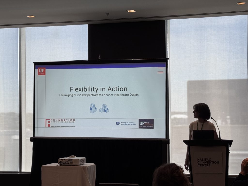 A woman stands at a podium in a conference room, presenting a slideshow titled Flexibility in Action: Leveraging Nurse Perspectives to Enhance Healthcare Design..
