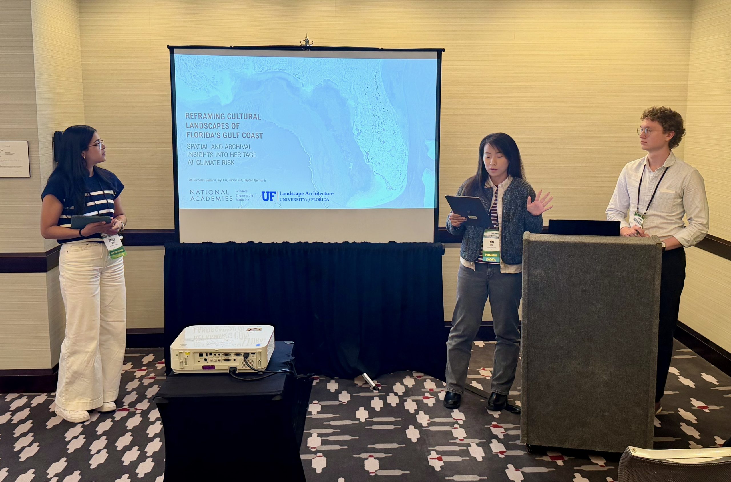 Three people stand in front of a projector screen in a conference room. The screen displays a presentation titled “Reframing Cultural Landscapes of Florida’s Gulf Coast.” One woman speaks while the others listen.