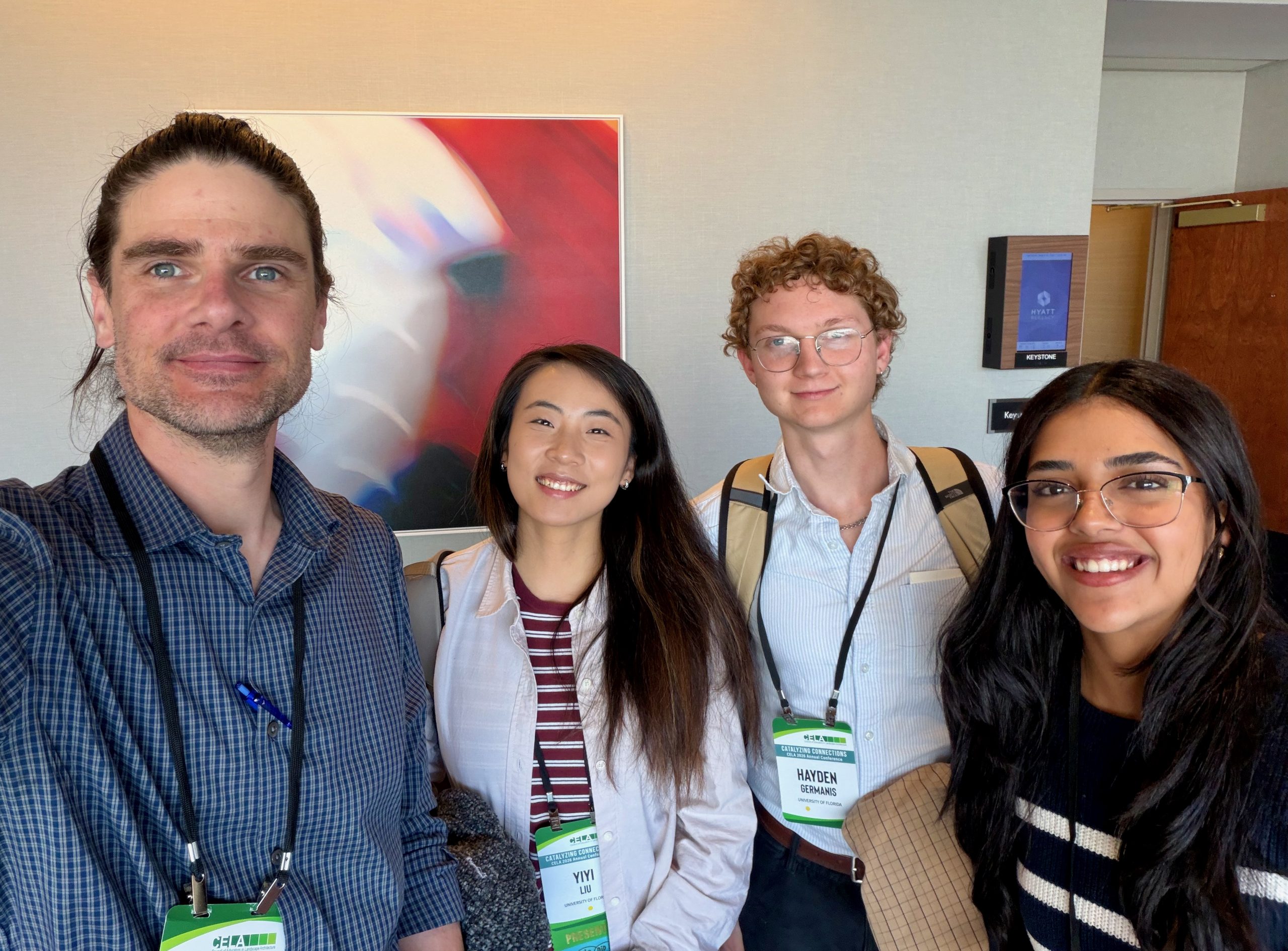 Four people wearing conference badges smile at the camera in a well-lit room with a blurred abstract painting on the wall behind them. Two men and two women are visible, all dressed in business casual attire.