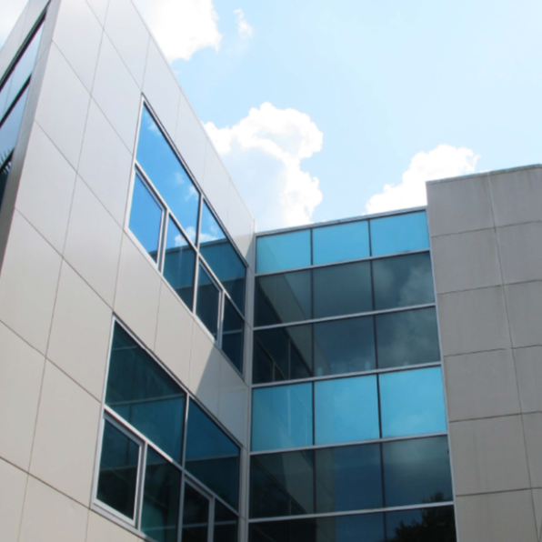 Modern building with large blue-tinted windows and light-colored walls, photographed from below against a blue sky with scattered clouds.
