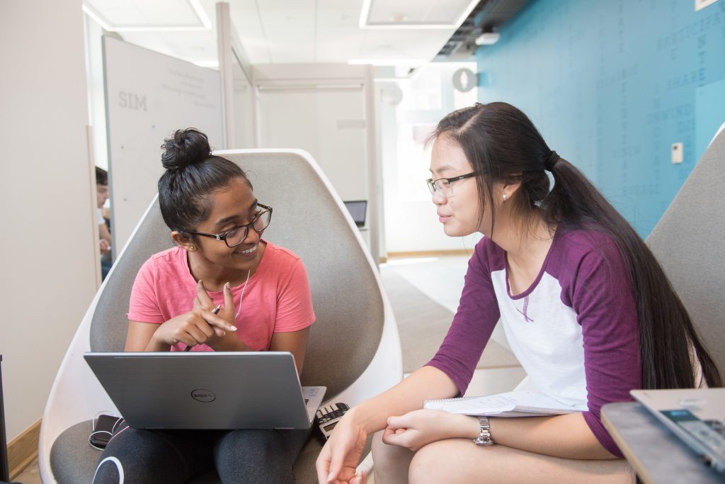 Two students studying and talking together in chairs with a computer.