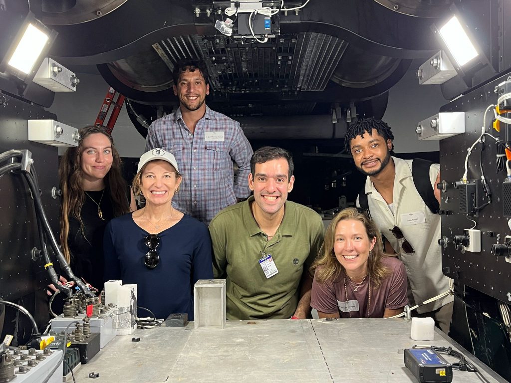 Mojtaba ‘Mo’ Tahmasebi is one of six people smiling and posing inside a high-tech, industrial space filled with metal equipment, cables, and various electronic components. Bright lights illuminate the scene from above and the sides.