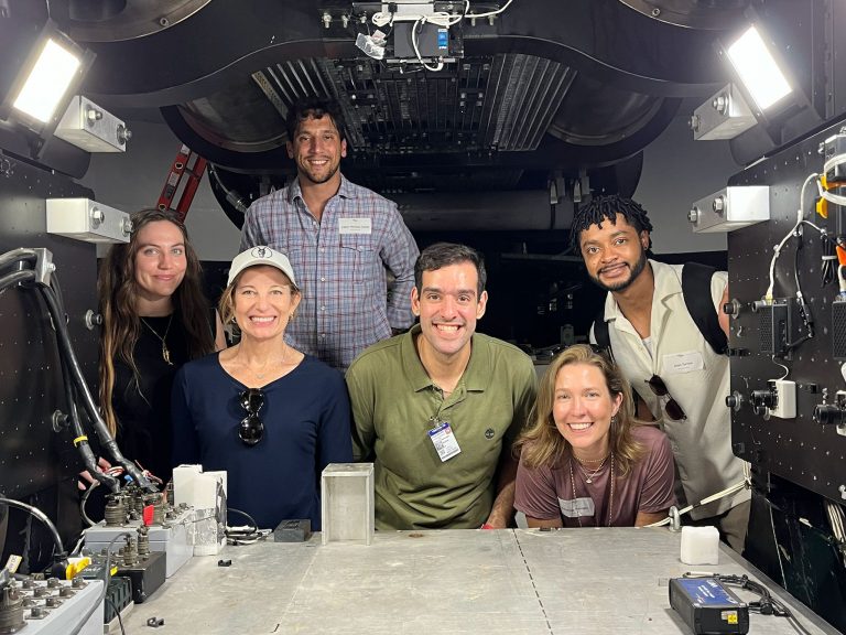 Six people smile and pose inside a high-tech, industrial space filled with metal equipment, cables, and various electronic components. Bright lights illuminate the scene from above and the sides.
