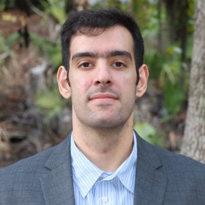 Mojtaba ‘Mo’ Tahmasebi with short dark hair wearing a gray suit jacket and a light blue striped shirt stands outdoors in front of trees and greenery, looking directly at the camera with a neutral expression.