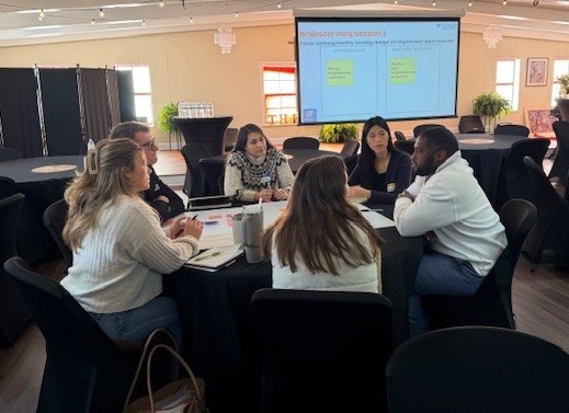 Six people sit around a table engaged in discussion with notebooks and laptops, in a large, well-lit room. A presentation slide about brainstorming is displayed on a screen behind them.