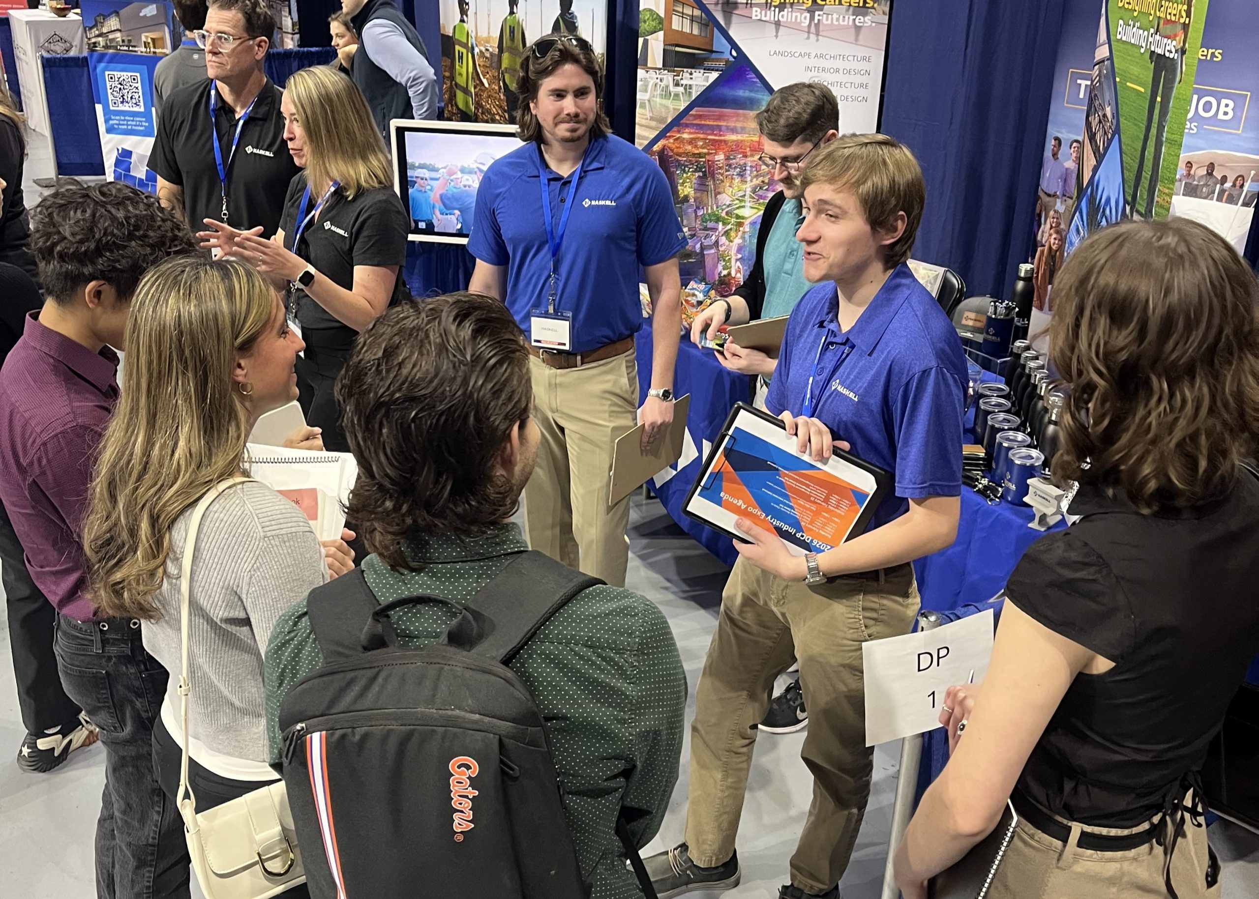 A group of students interact with recruiters at a career fair booth. One recruiter in a blue shirt holds a tablet and speaks to attendees while others listen or look at flyers. The booth has a blue tablecloth and promotional displays.