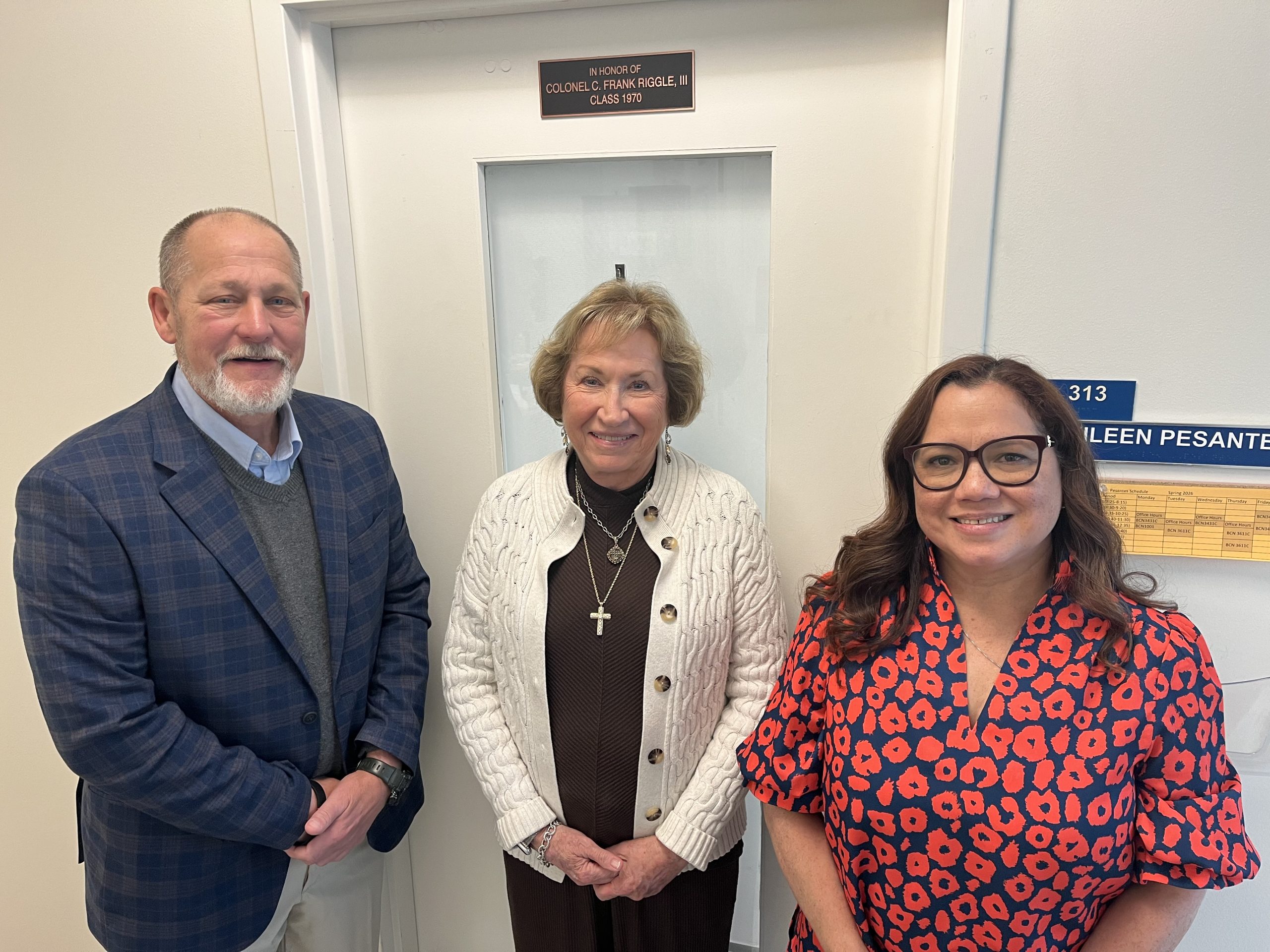 Three adults stand smiling in front of a white door. The person on the left wears a blue plaid jacket, the person in the center wears a white sweater, and the person on the right wears glasses and a red patterned dress.