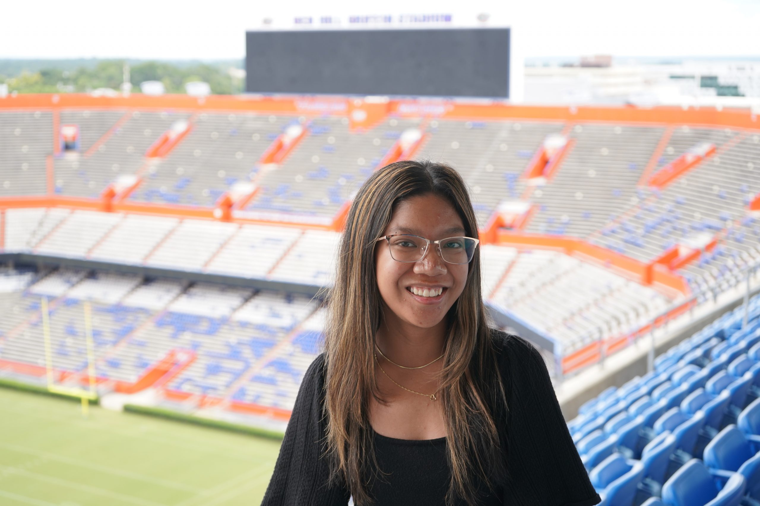 Hope Castillo smiles while sitting in the stands of a large, mostly empty football stadium with blue and orange seats.