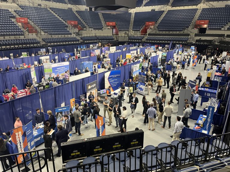 A large indoor career fair with rows of booths, recruiters at tables, and many attendees walking and talking. The event is held in an arena with blue and gray seating in the background.