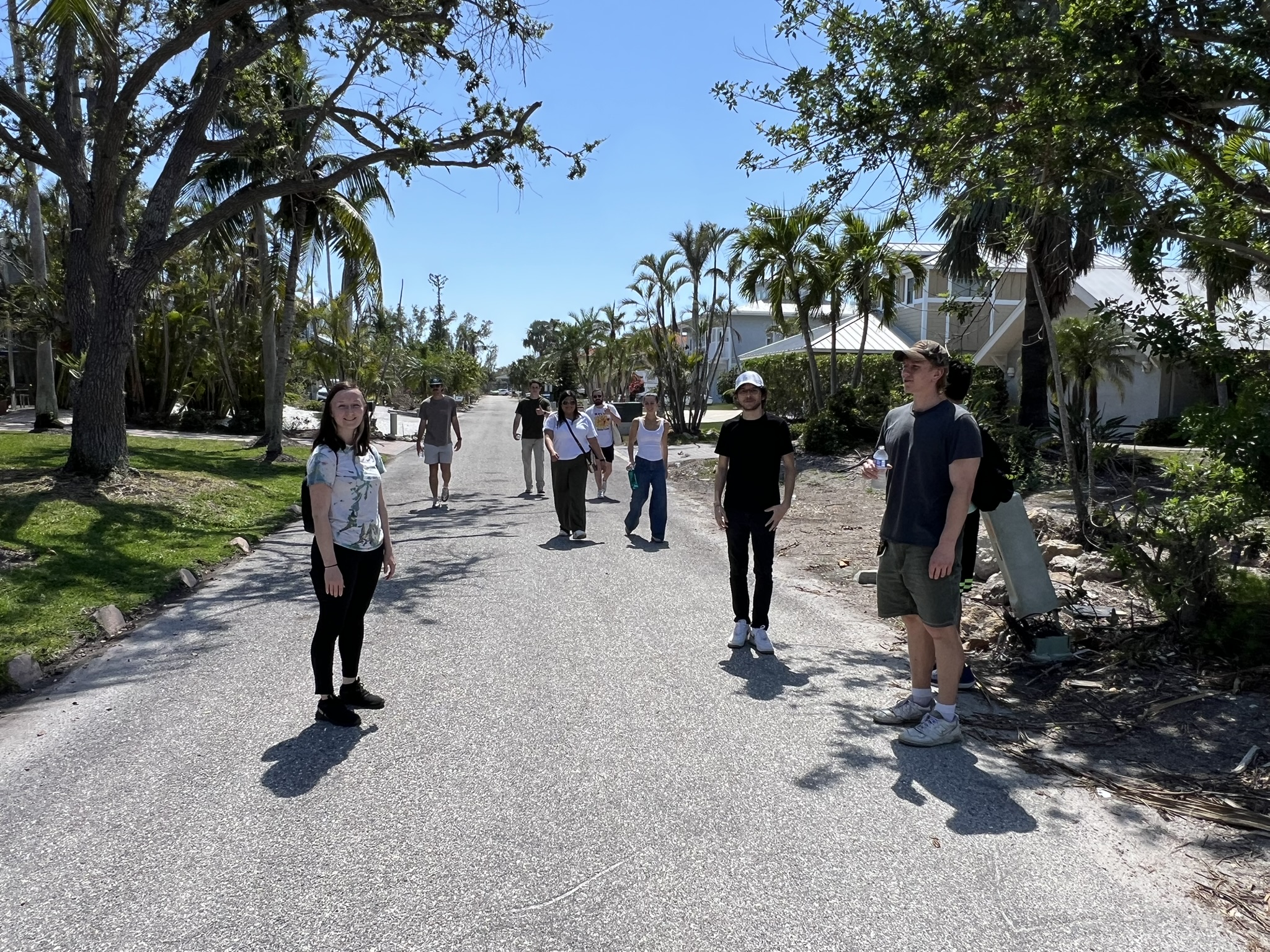 A group of people stands spaced out along a sunlit residential street lined with trees and houses. Some stand in pairs while others are alone, and shadows are cast on the pavement. The weather is clear and sunny.