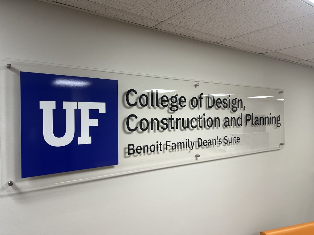 A wall sign with a blue UF logo and text reading College of Design, Construction and Planning, Benoit Family Deans Suite on a clear panel.