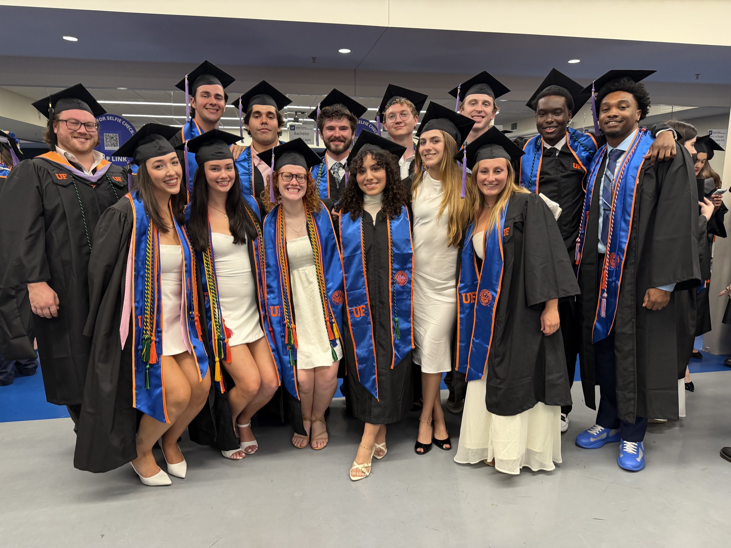 A group of graduates in caps and gowns, many wearing blue and orange stoles, pose together indoors. Several are smiling with arms around each other. Most are dressed in white outfits under their gowns.