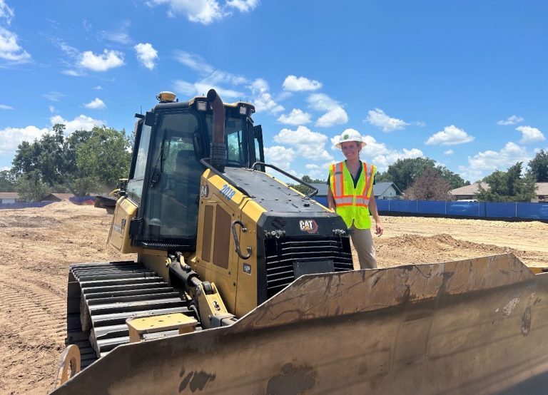 A person in a white hard hat and neon safety vest stands next to a yellow CAT bulldozer at a construction site under a bright blue sky with scattered clouds.