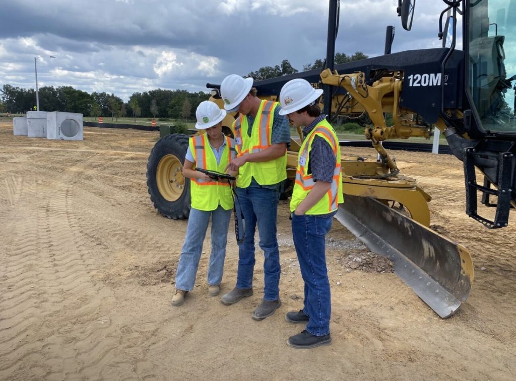 Three construction workers in safety vests and hard hats stand on a dirt construction site, looking at a tablet together beside a large yellow grader on a cloudy day.