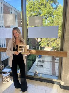 A woman stands indoors holding a small architectural model. Behind her, large windows display tree-filled views and sketches are taped to the glass. Sunlight streams in, lighting the scene.