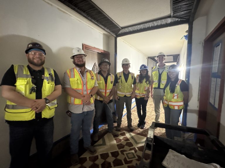 Seven people wearing hard hats and safety vests stand in a hallway under construction, smiling at the camera. The hallway has exposed wiring and a patterned tile floor.
