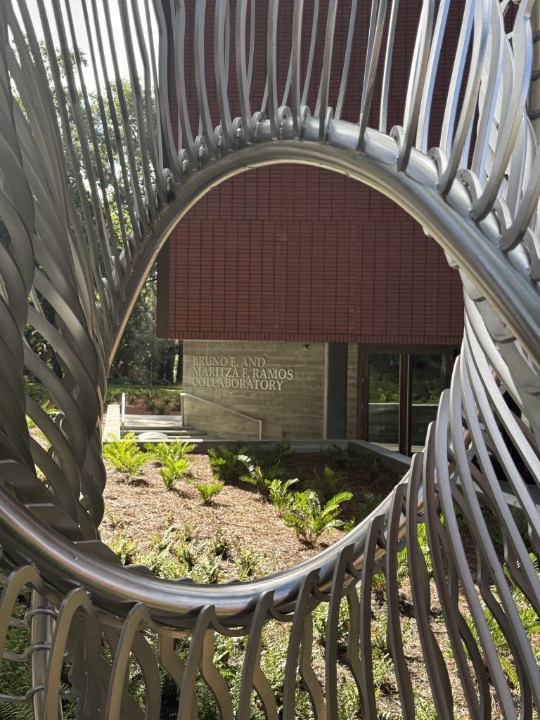 View through a circular metal sculpture, framing a building with a sign that reads Bruno E. and Reitza A. Ramos Collaboratory. Greenery and sunlight are visible in the scene.