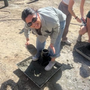 A smiling woman wearing sunglasses pours concrete into a metal cone on a tray outdoors, with other people working nearby in the sunlight.