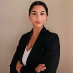 A woman with dark hair tied back, wearing a black blazer and white top, stands with arms crossed and smiles confidently in front of a plain beige wall.