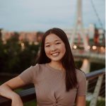 A young woman with long brown hair smiles while standing on a bridge with city lights and a large structure in the background at dusk.