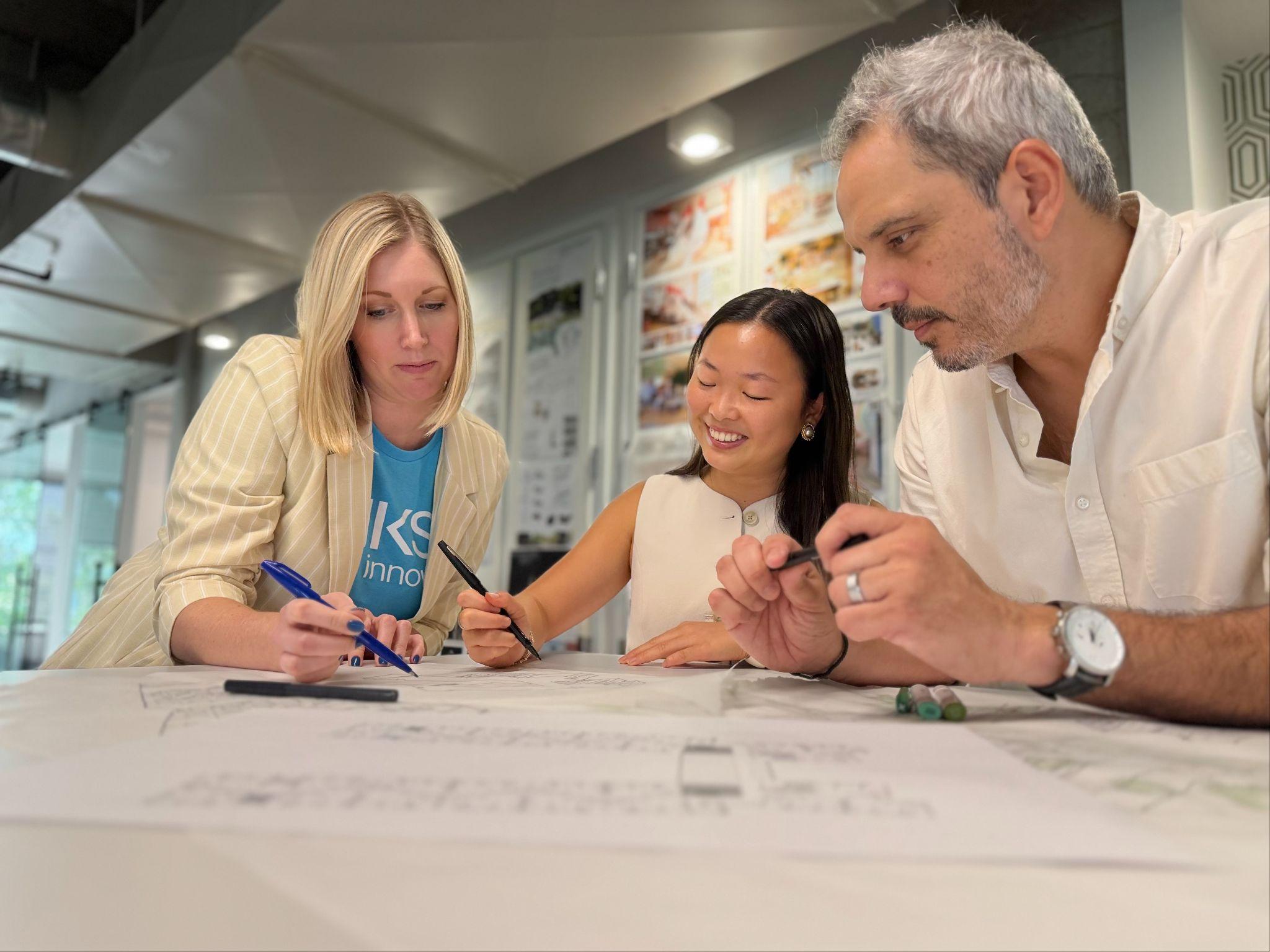Three people sit at a table, collaborating on architectural drawings. One woman writes with a pen, another woman smiles, and a man looks focused, all working together in a modern office setting.