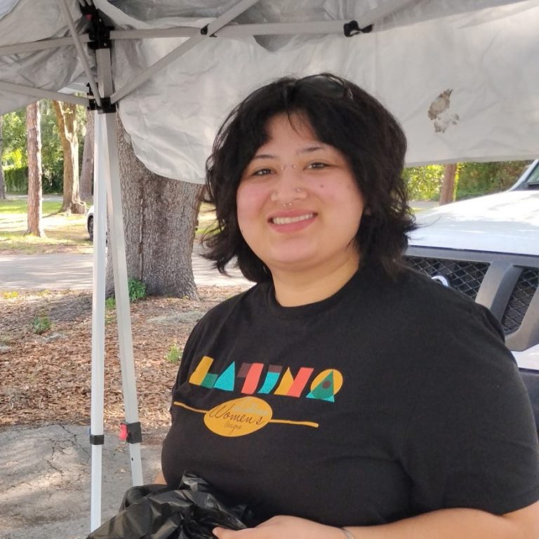 A person with short dark hair smiles under a white canopy outdoors, wearing a black Latina Women t-shirt. Trees and a white vehicle are visible in the background.