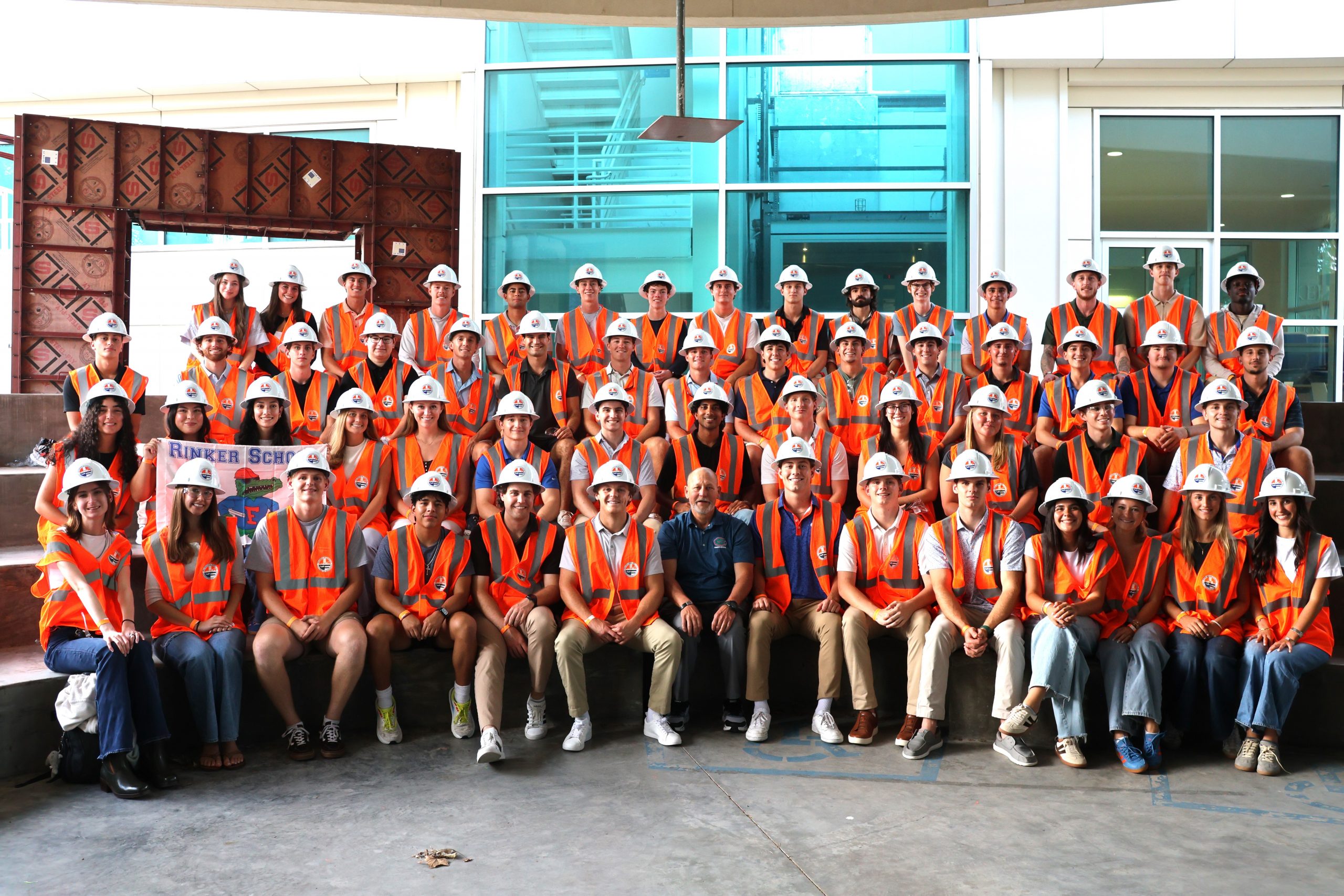 A large group of people in orange safety vests and white hard hats pose for a photo indoors; some are seated while others stand behind them.