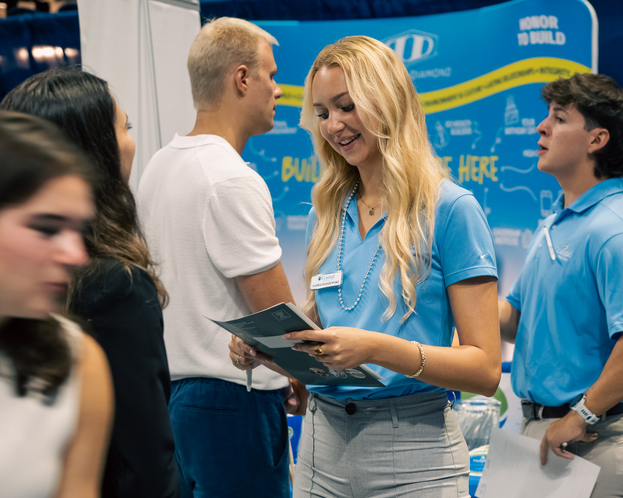 A woman in a blue shirt smiles while holding papers at a busy career fair booth. Several people are talking in the background. Blue banners and informational displays are visible behind her.