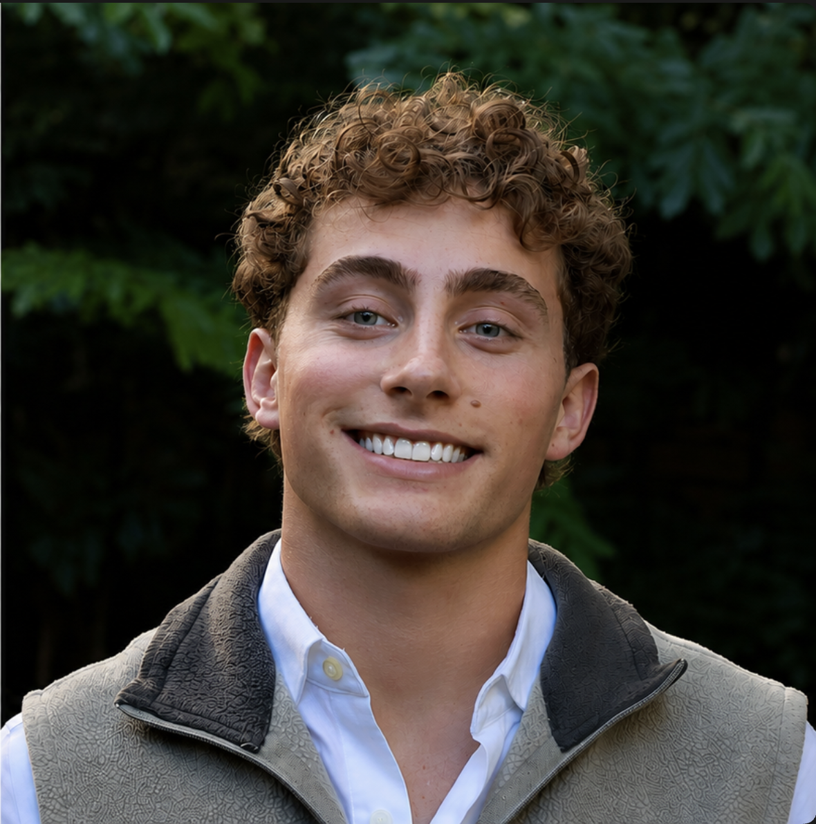 A young man with curly brown hair smiles outdoors. He is wearing a white shirt and a gray fleece vest, with green foliage in the background.