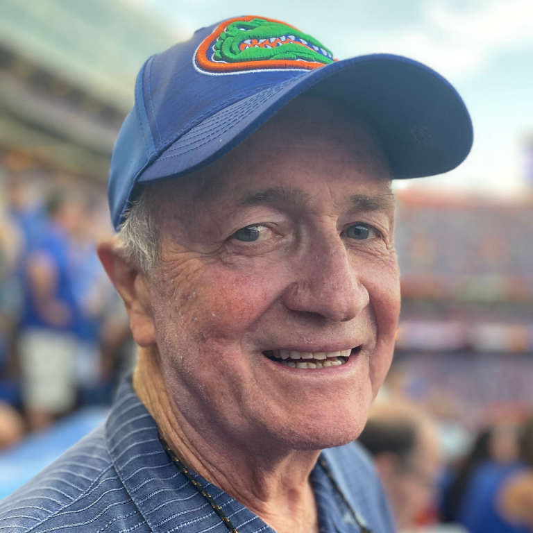 Older man wearing a blue Florida Gators baseball cap and striped shirt smiles at a sports stadium, with blurred fans and seating in the background.