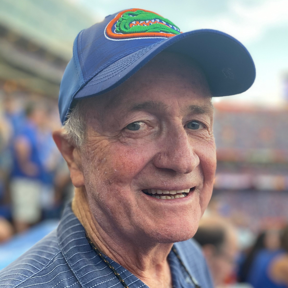 Older man wearing a blue Florida Gators baseball cap and striped shirt smiles at a sports stadium, with blurred fans and seating in the background.