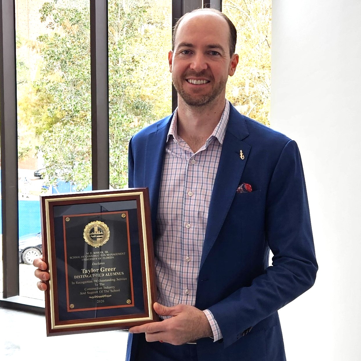 A man in a blue blazer and plaid shirt holding a plaque that reads, Taylor Greer, Distinguished Alumnus, In Recognition Of Outstanding Service To The Construction Industry And Support Of The School.